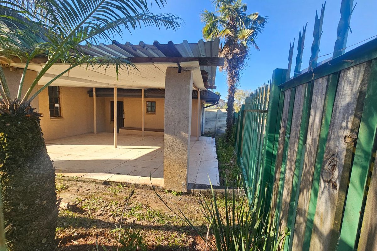 A covered outdoor patio area featuring tiled flooring and concrete support pillars, adjacent to a metal perimeter fence.