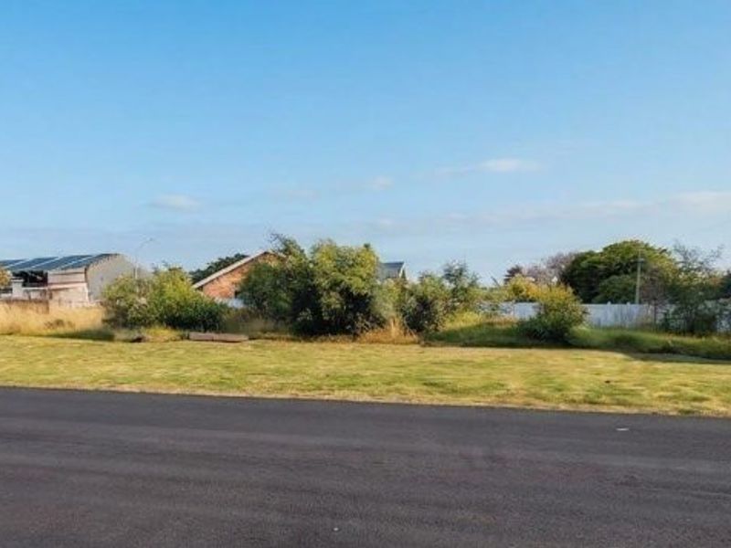 A vacant residential plot featuring a level grassed surface, bordered by precast concrete walls and accessible via a paved road.