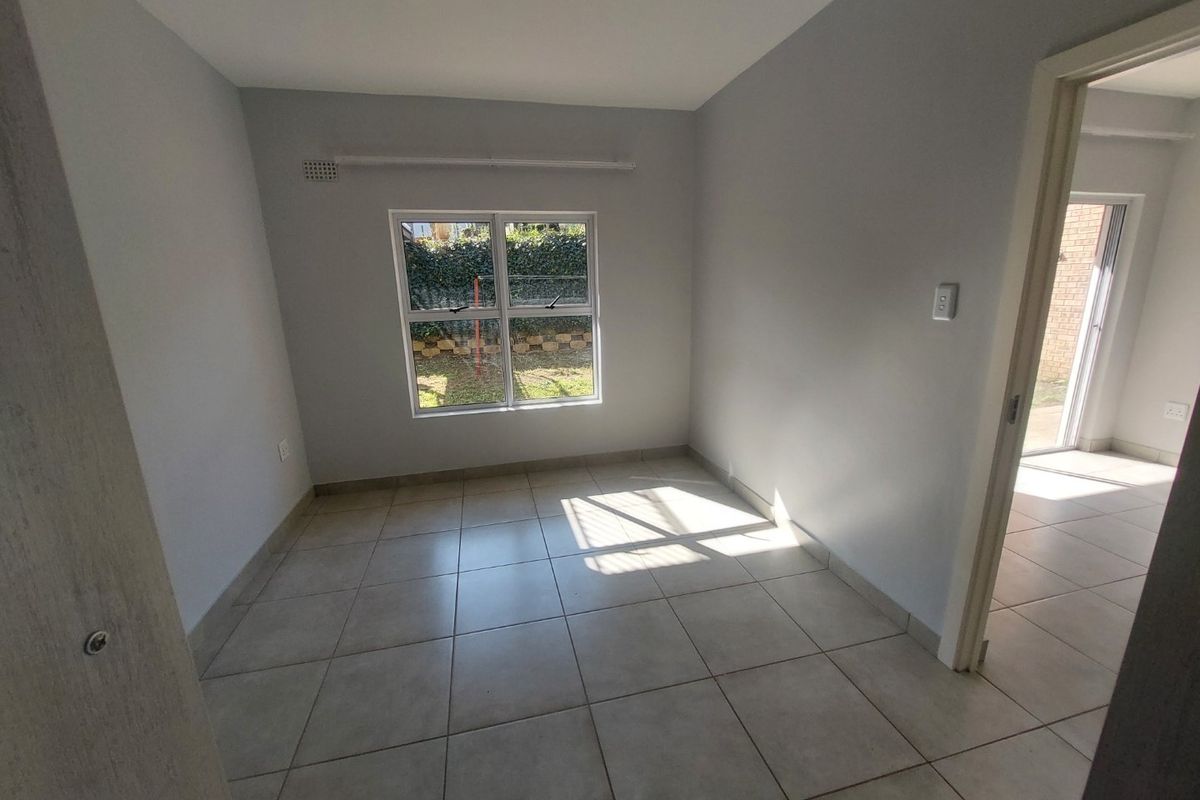 A bright, empty bedroom featuring neutral tiled flooring, clean walls, and a window overlooking the garden area.
