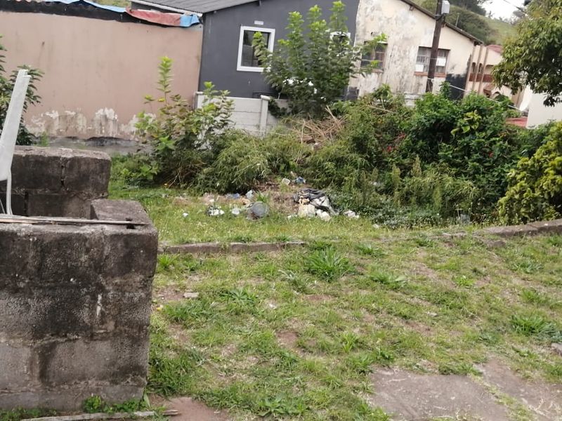 A neglected backyard area featuring overgrown grass, scattered debris, and a weathered concrete boundary wall requiring significant maintenance and landscaping.