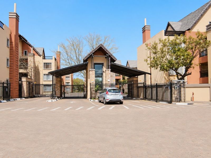 A well-maintained apartment complex entrance featuring a secure gatehouse, paved driveway, and multi-story residential buildings with balconies.