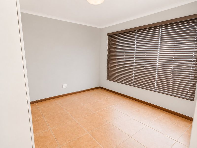 A clean, empty bedroom featuring neutral wall paint, tiled flooring, and fitted venetian blinds, offering a versatile space ready for personalization.