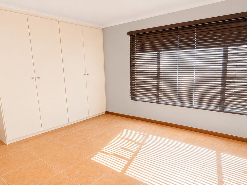 A bright, empty bedroom featuring neutral tiled flooring, built-in cupboards, and dark venetian blinds on the window.