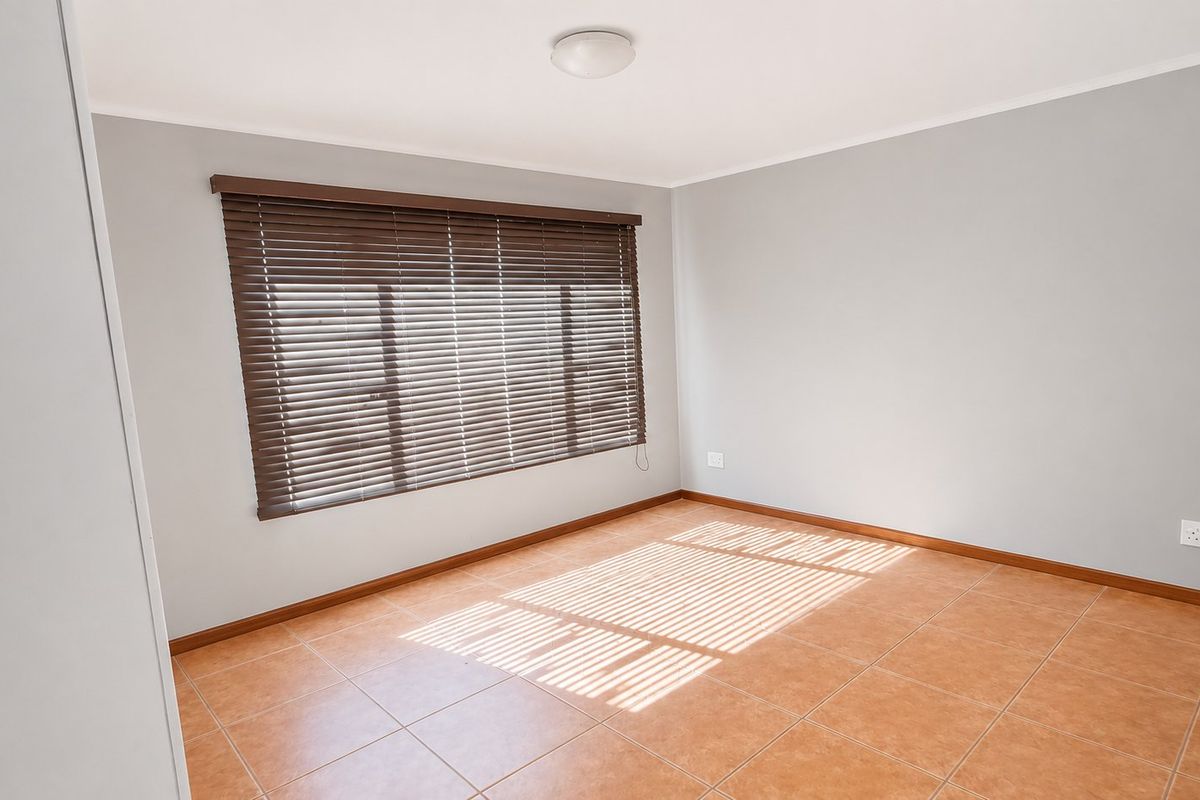 A bright, empty bedroom featuring neutral grey walls, terracotta-style floor tiles, and fitted venetian blinds.
