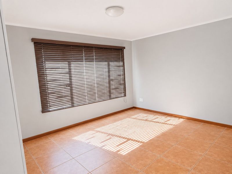 A bright, empty bedroom featuring neutral grey walls, terracotta-style floor tiles, and fitted venetian blinds.