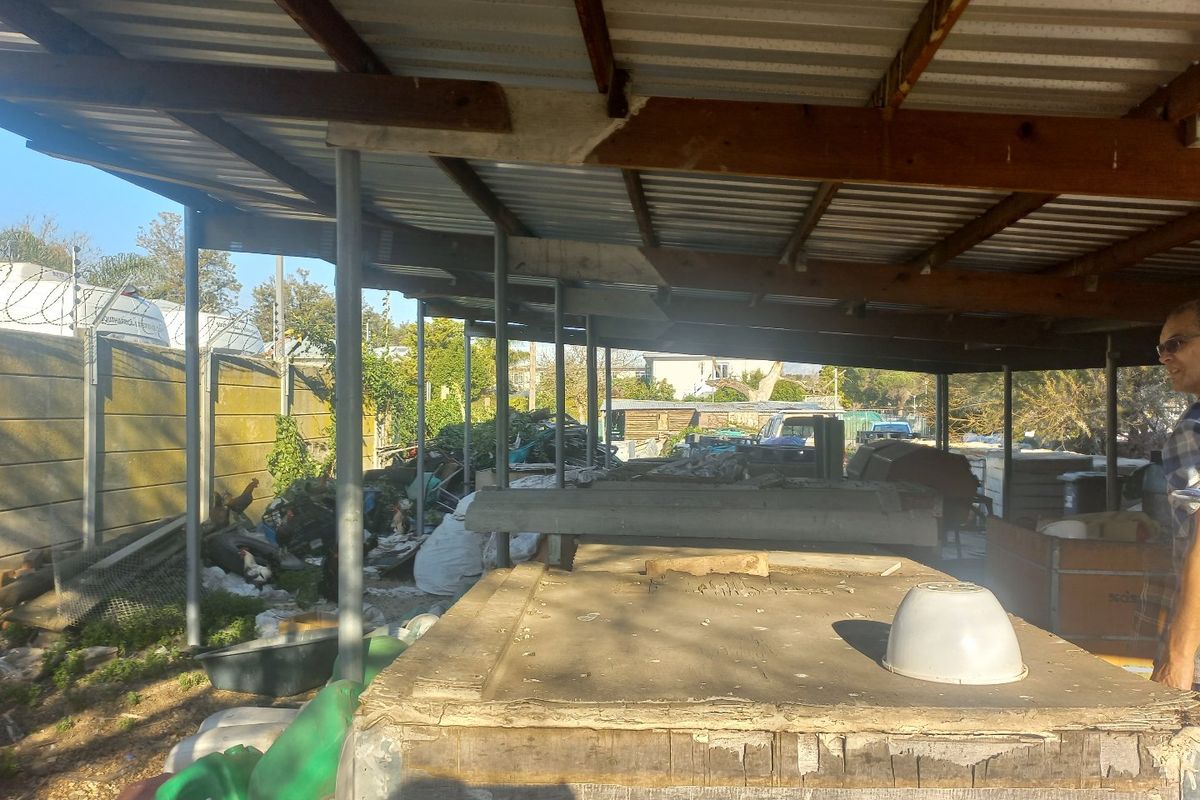 A covered outdoor utility or storage area featuring a corrugated metal roof supported by steel posts and a precast concrete boundary wall.