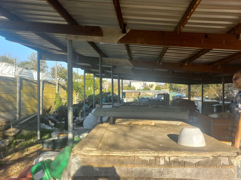 A covered outdoor utility or storage area featuring a corrugated metal roof supported by steel posts and a precast concrete boundary wall.