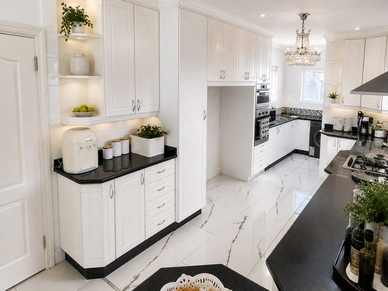 A modern, well-appointed kitchen featuring white cabinetry, contrasting black granite countertops, high-end built-in appliances, and elegant marble-look flooring with decorative crown molding.