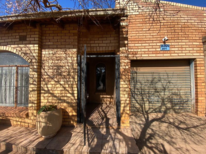 Front elevation of a face brick house featuring a secure entrance gate, tiled porch area, and a roller shutter garage door.