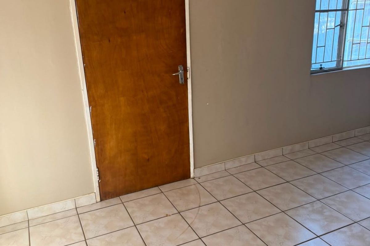 An empty bedroom featuring neutral ceramic floor tiles, a wooden door, and a window fitted with security bars.