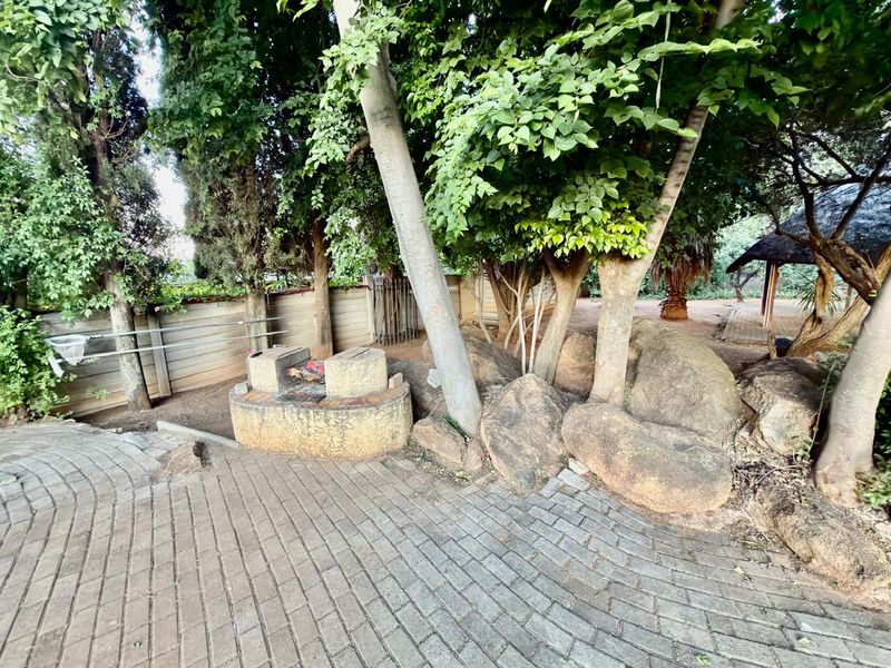 A rustic garden area featuring a built-in stone braai, paved patio flooring, and mature trees, offering a natural outdoor entertainment space.