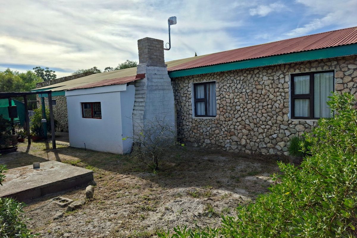 Rear view of a rustic stone-clad house featuring a prominent brick chimney, corrugated metal roofing, and aluminum-framed windows set in a natural garden area.