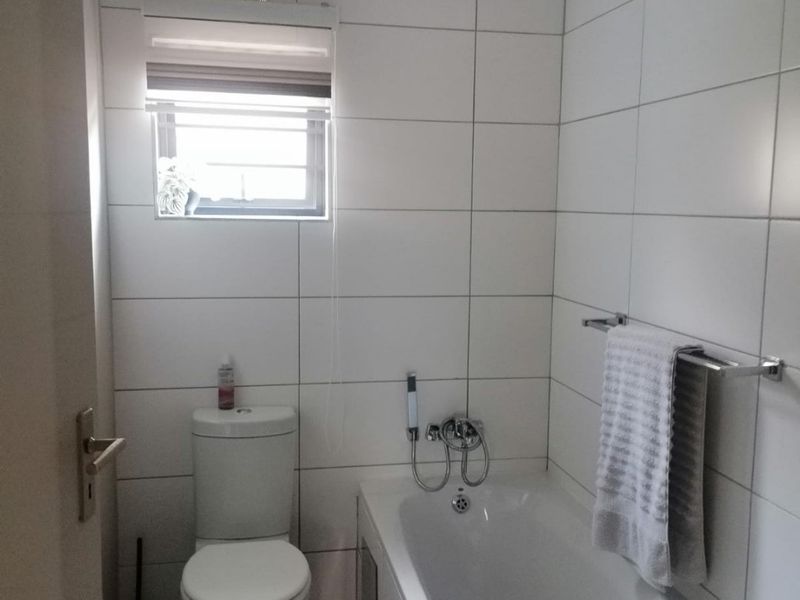 A clean, modern bathroom featuring white wall tiles, a standard bathtub, and a toilet, with a small window providing natural light.