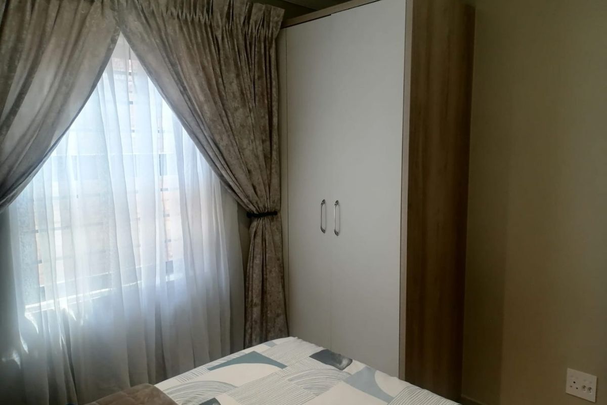 A bedroom featuring a modern built-in wardrobe with white doors and wood-grain side paneling, positioned next to a window with draped curtains.