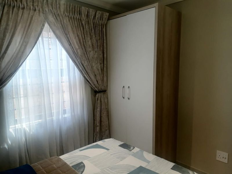 A bedroom featuring a modern built-in wardrobe with white doors and wood-grain side paneling, positioned next to a window with draped curtains.