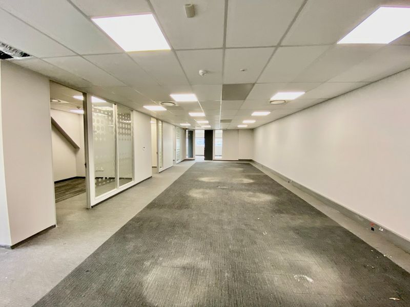 An empty commercial office space featuring glass partitions, suspended ceilings with recessed lighting, and worn carpet flooring throughout.