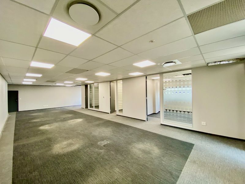 An open-plan commercial office space featuring suspended ceilings, recessed lighting, and glass-partitioned private offices with worn carpeting throughout.