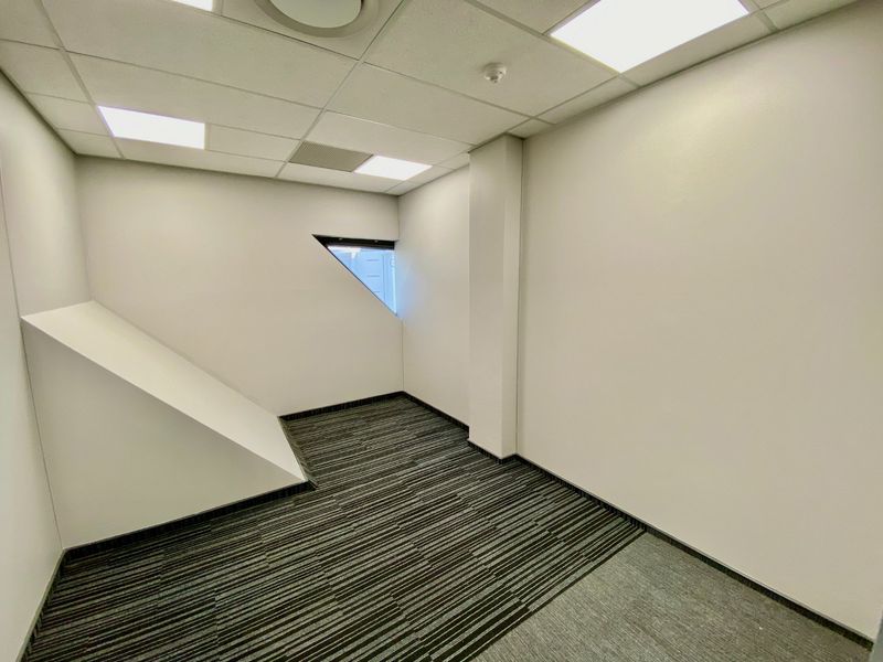 A clean, empty commercial office space featuring modern carpet tiles, a suspended ceiling with recessed lighting, and a unique triangular window.