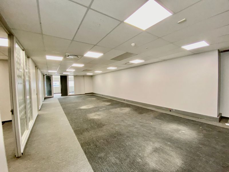 An empty, open-plan commercial office space featuring a suspended ceiling with recessed lighting, glass partitions, and carpeted flooring.
