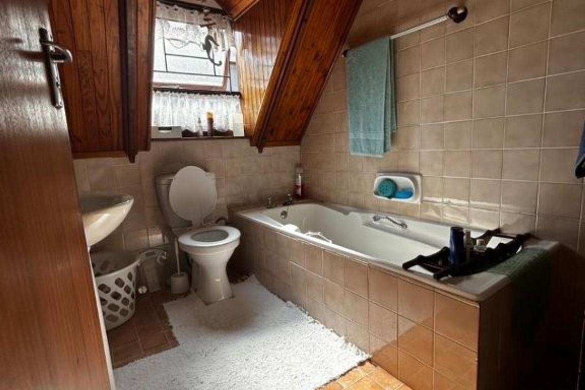 A dated bathroom featuring a built-in bathtub, pedestal sink, toilet, and distinctive timber-paneled vaulted ceiling with tiled finishes.