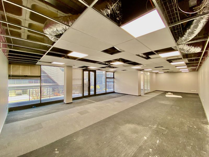 Open-plan commercial office space featuring large windows, carpeted flooring, and a partially finished suspended ceiling with exposed ducting.