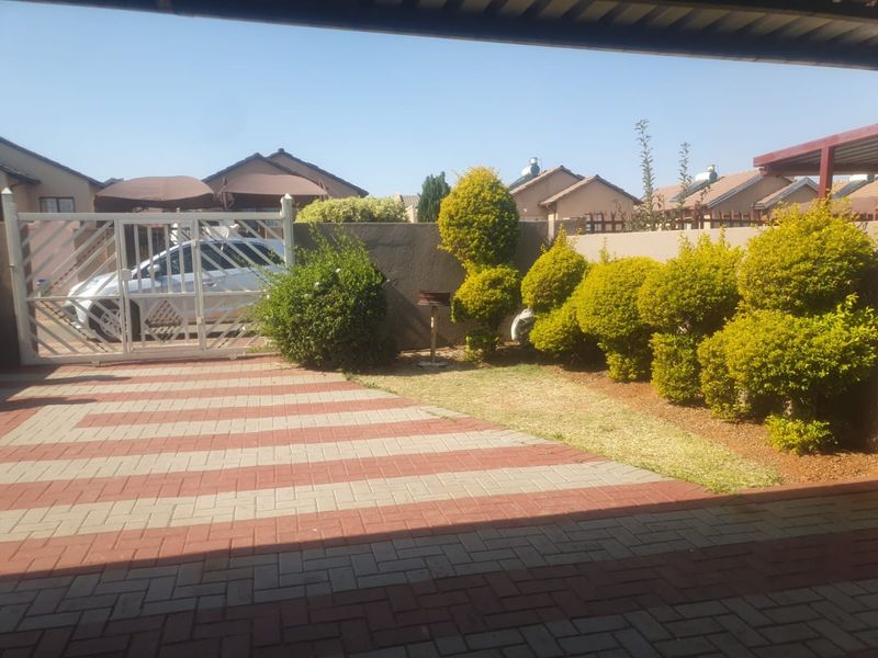 A neat front exterior view featuring a paved driveway, secure metal gate, and established garden landscaping with a boundary wall.
