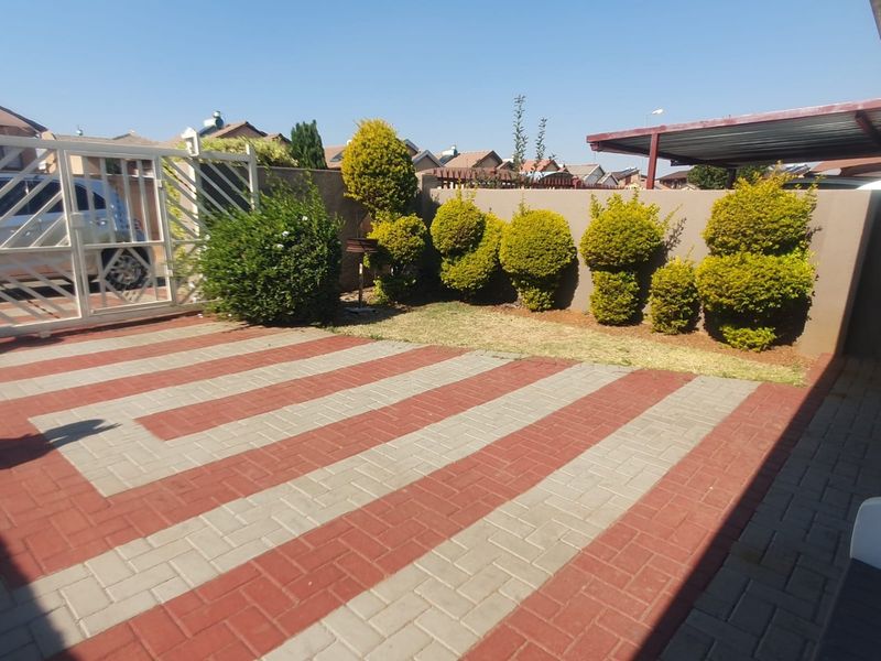 A paved front driveway featuring a patterned brick design, enclosed by a boundary wall with a metal gate and an adjacent carport structure.