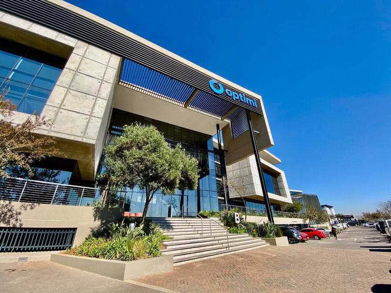 Modern commercial office building featuring a prominent glass facade, concrete architectural elements, and a paved entrance area with professional landscaping.