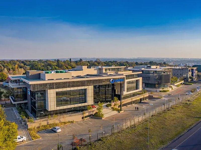 Aerial view of a modern multi-story commercial office building featuring glass facades, extensive parking facilities, and landscaped surroundings in a business park setting.