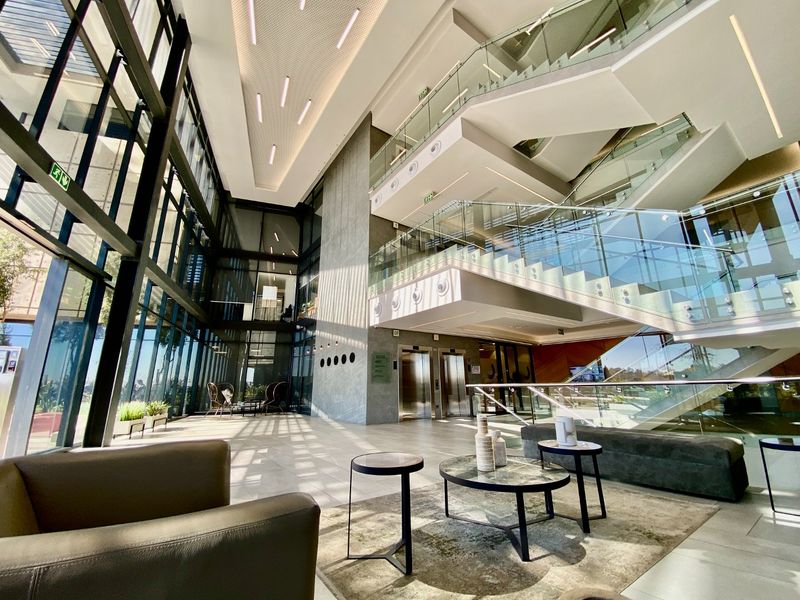 Modern commercial lobby featuring a dramatic double-volume atrium, floor-to-ceiling glass facade, floating staircase with glass balustrades, and high-end finishes.