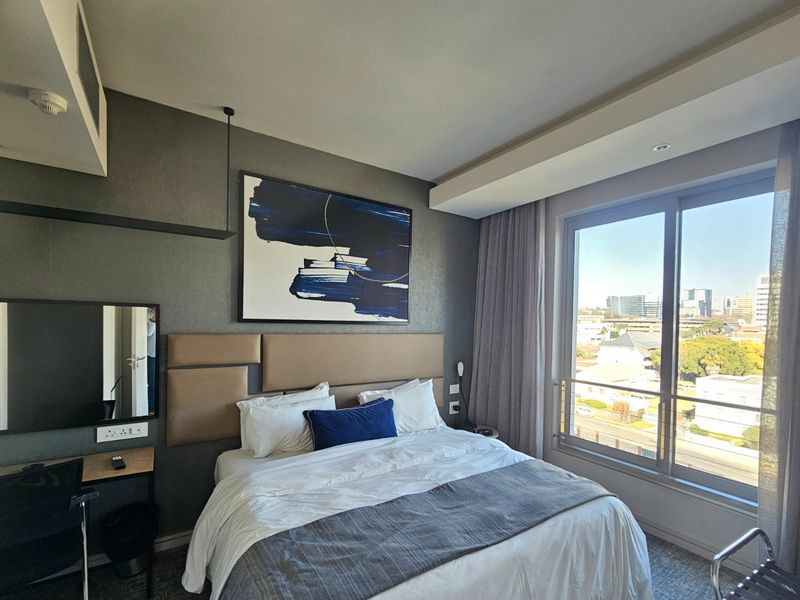 Modern bedroom featuring a neutral upholstered headboard, built-in desk area, and large sliding glass doors offering expansive city views.