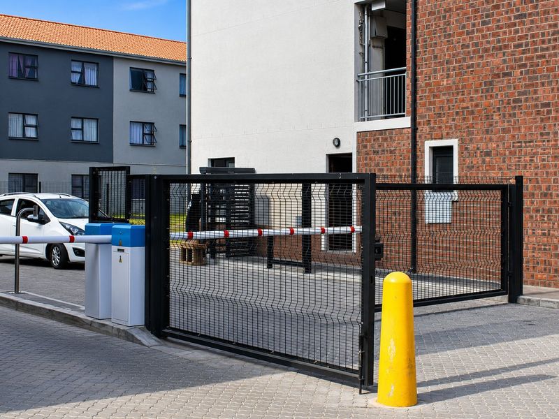 A secure apartment complex entrance featuring a boom gate, mesh security fencing, and paved driveway access.