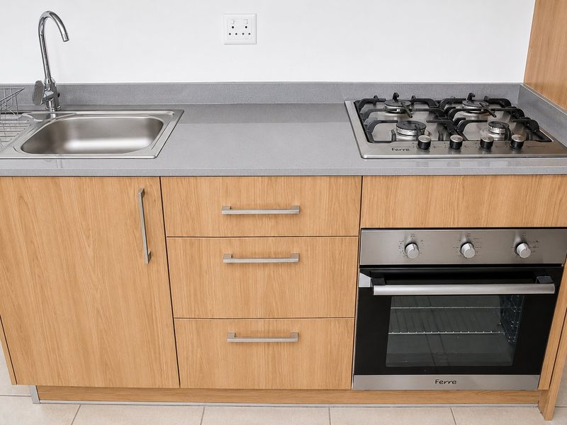 Modern kitchen kitchenette featuring light wood cabinetry, stone countertops, a stainless steel sink, and a built-in gas hob with oven.