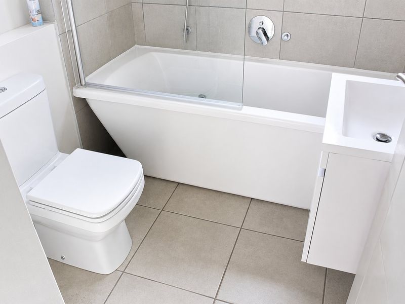 A modern, compact bathroom featuring a bathtub with a glass screen, a wall-mounted vanity basin, and a toilet, finished with neutral floor and wall tiles.