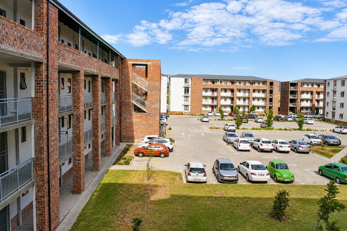 An elevated view of an apartment complex featuring exposed brick architecture, metal balcony railings, and a paved parking lot with landscaped green spaces.