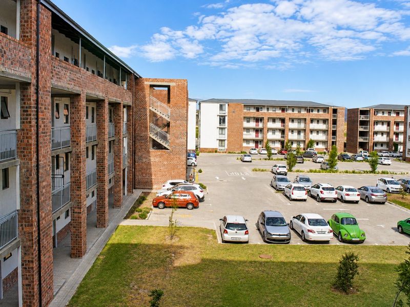 An elevated view of an apartment complex featuring exposed brick architecture, metal balcony railings, and a paved parking lot with landscaped green spaces.