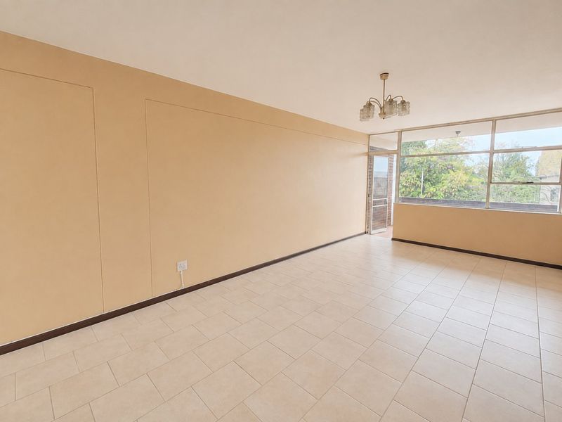 A spacious, empty living area featuring neutral tiled flooring, large windows providing natural light, and access to an outdoor balcony area.
