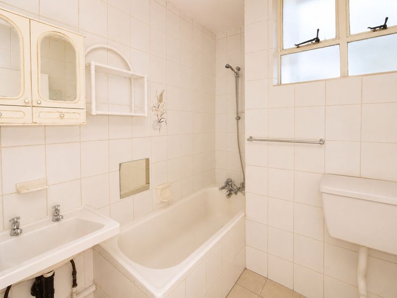A dated bathroom featuring a bathtub, wall-mounted basin, and toilet, finished with white wall tiles and neutral floor tiles.