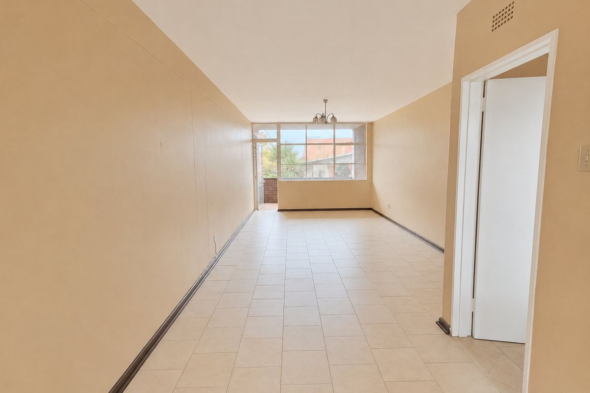 A spacious, empty living area featuring neutral tiled flooring, large windows providing natural light, and access to an outdoor balcony.
