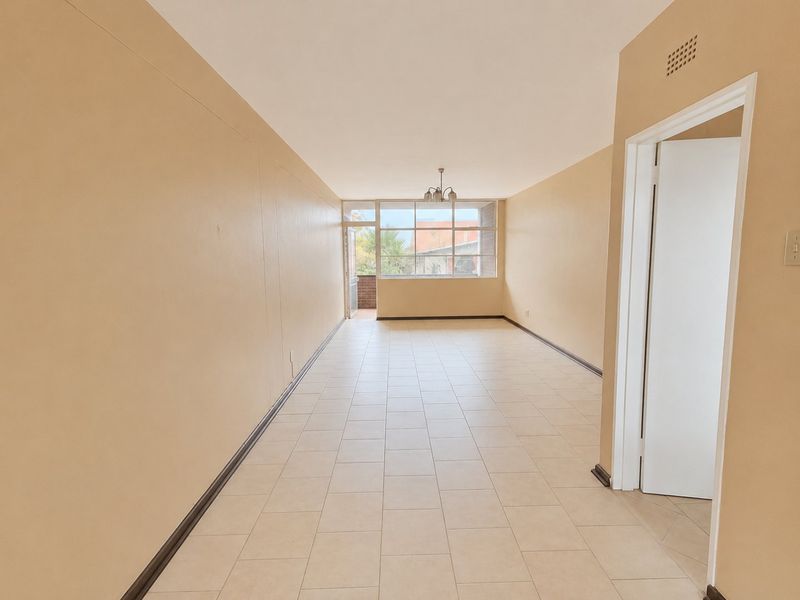 A spacious, empty living area featuring neutral tiled flooring, large windows providing natural light, and access to an outdoor balcony.