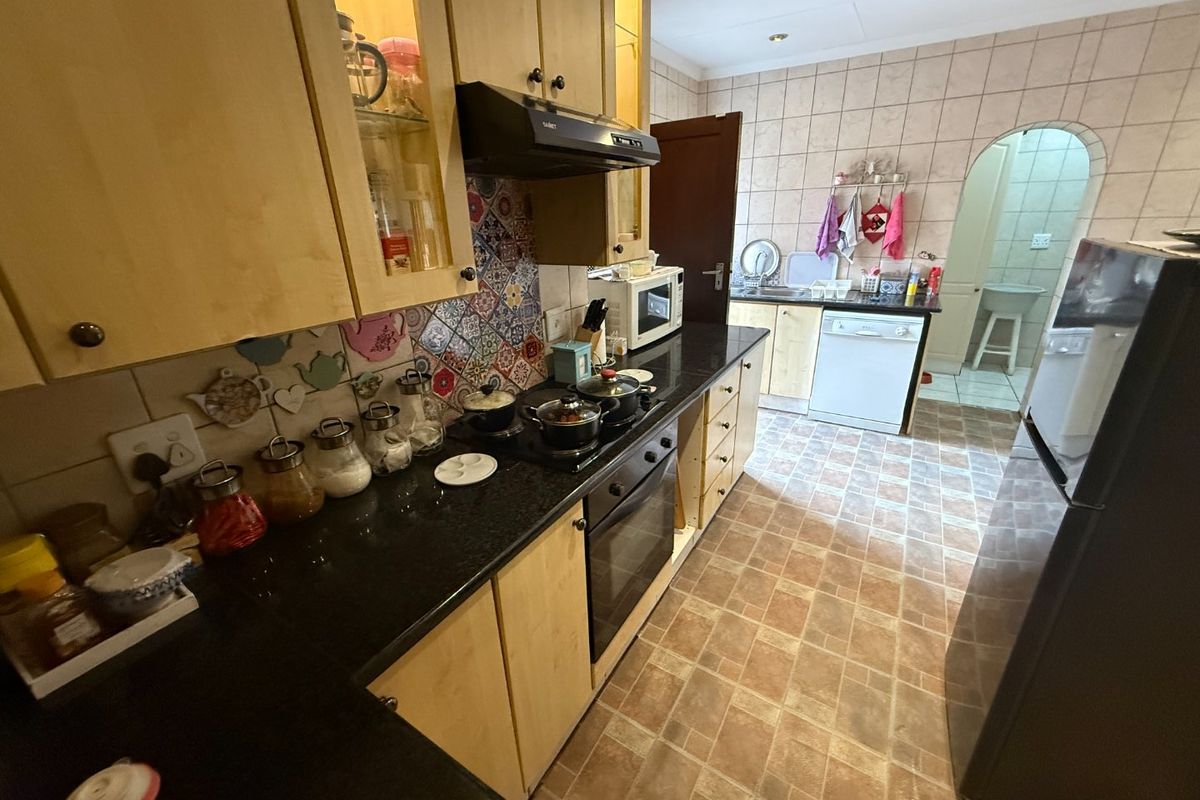 A functional kitchen featuring light wood cabinetry, dark granite countertops, a built-in oven with hob, and a tiled backsplash, leading into a separate scullery area.