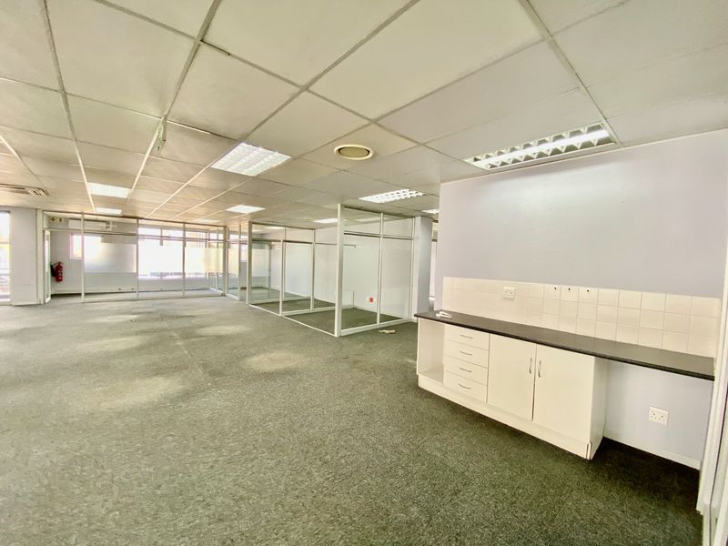 Open-plan commercial office space featuring glass partitions, a small kitchenette area, suspended ceilings, and carpeted flooring throughout.