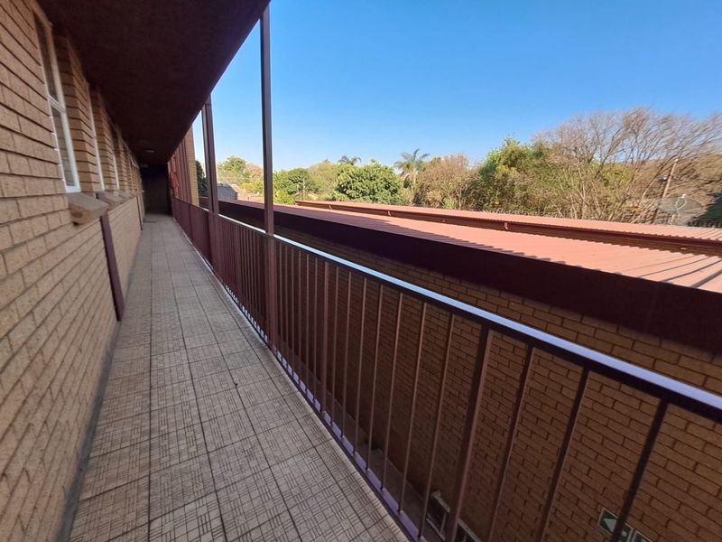 An exterior covered balcony walkway featuring brick walls, tiled flooring, and a metal safety railing overlooking the surrounding area.