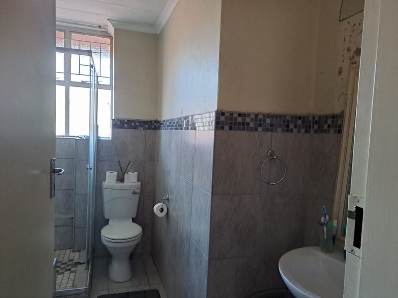 A functional bathroom featuring a shower enclosure, toilet, and pedestal basin, with tiled walls and visible wall surface damage requiring repair.