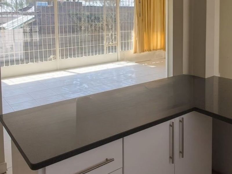 A clean, modern kitchen island with white cabinetry and dark countertops, overlooking an adjacent tiled patio area through a large opening.