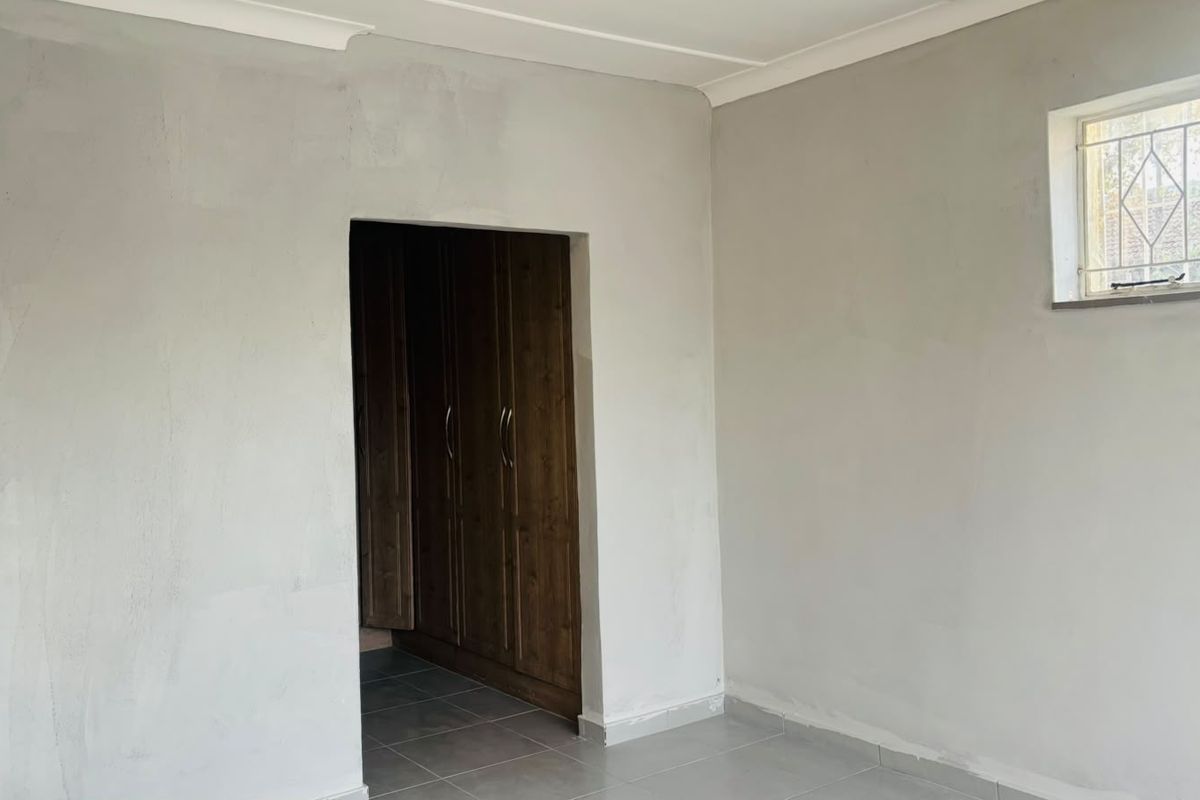 An empty bedroom featuring tiled flooring, built-in wooden cupboards, and security bars on the window, requiring minor cosmetic finishing to the walls.