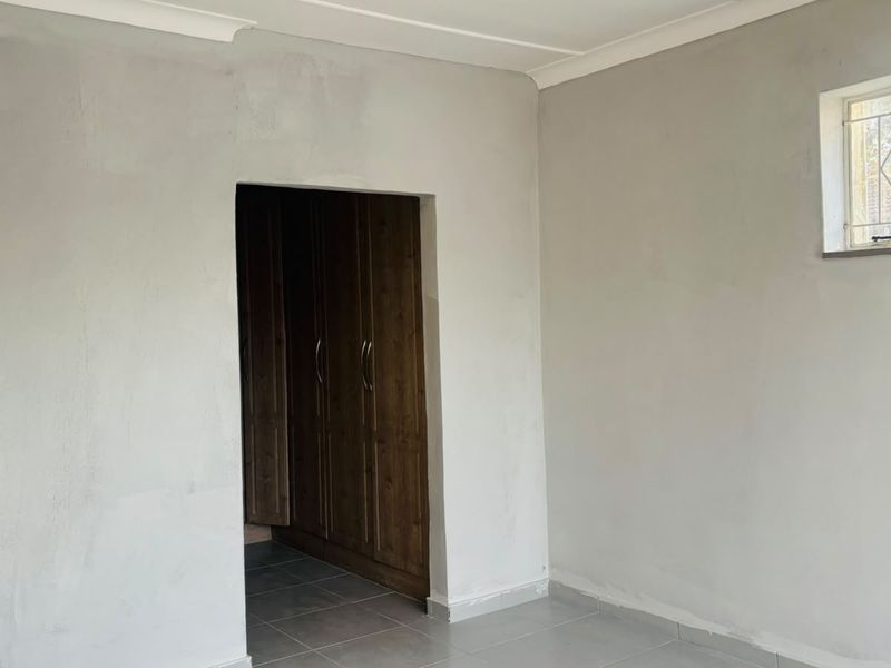 An empty bedroom featuring tiled flooring, built-in wooden cupboards, and security bars on the window, requiring minor cosmetic finishing to the walls.