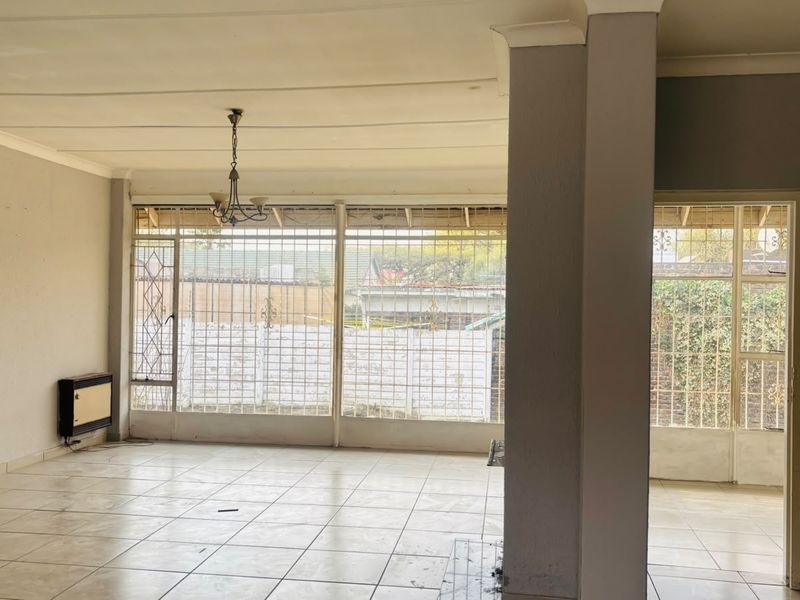 An empty, open-plan living area featuring tiled flooring, security bars on windows, and a wall-mounted heater, offering a blank canvas for renovation.