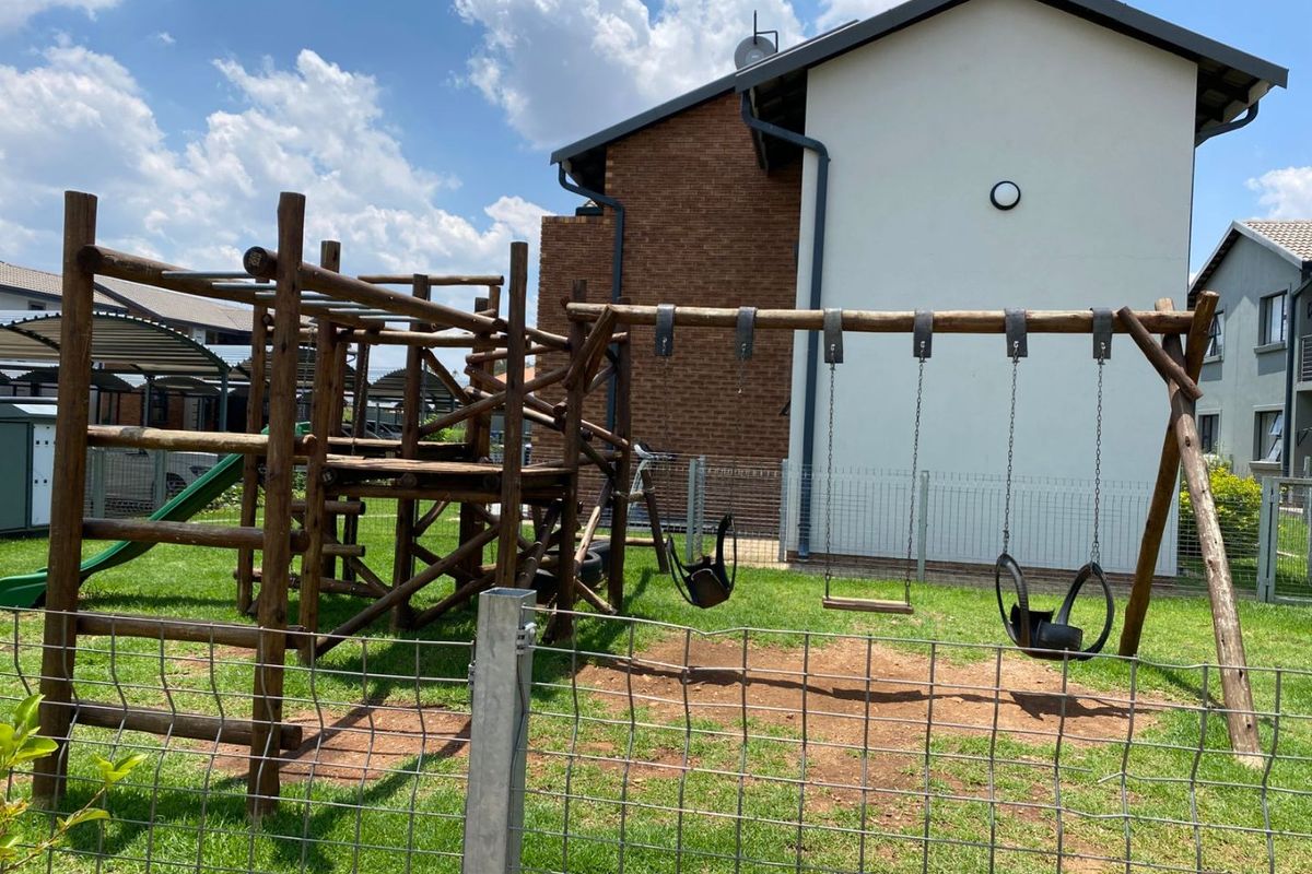 A communal outdoor area featuring a wooden playground structure with swings, set within a fenced lawn area adjacent to the residential building.