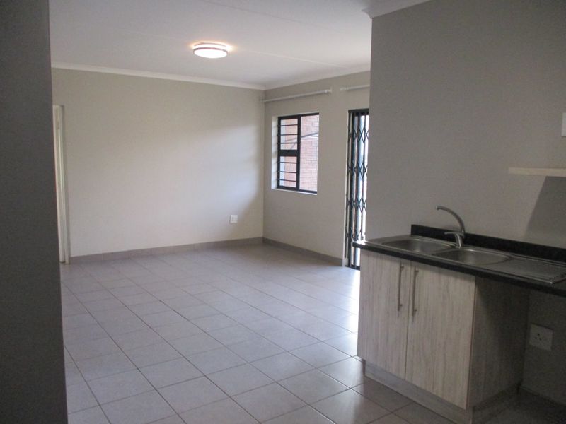 An open-plan living and kitchen area featuring neutral tiled flooring, built-in kitchen cabinetry with a sink, and access to an exterior security gate.